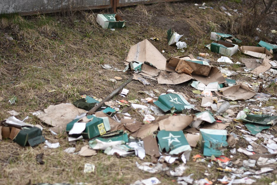 An outdoor scene showing a patch of ground strewn with discarded packaging materials, including crushed and torn cardboard boxes with a green and white logo, and scattered paper debris. The boxes appear to be from food or beverage products, with some flattened or partially collapsed, revealing their corrugated textures. The debris is spread across a grassy area adjacent to a paved driveway, with a slight incline leading up to a rusty metal fence in the background. Among the litter, there are small pieces of torn paper, plastic wrappers, and bits of cardboard, creating a cluttered and unmanaged appearance. The environment is naturally lit, with an overcast sky casting diffuse light over the scene, highlighting the uneven surface and the various textures of the trash. This image visually illustrates issues related to illegal dumping or inadequate waste collection, emphasizing the need for professional rubbish removal services such as those offered by Rubbish Removal Harrow, to maintain clean and safe outdoor spaces and facilitate proper waste handling and disposal alternatives to roadside dumping.