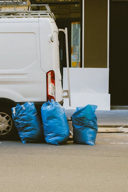 The image shows the rear side of a white delivery van parked on a paved street in front of a building with large windows. Three blue trash bags, made of plastic with slightly crinkled textures, are positioned on the ground near the occupancy side of the van, suggesting they are set aside for rubbish removal or private waste disposal. The bags are filled and tied at the top, with the middle bag leaning slightly to the left. Behind the van, part of the building’s exterior wall is visible, including a white railing or panel at ground level and a glass door or window reflecting the environment. The scene appears to be outdoors during daylight, with natural lighting illuminating the area. This configuration of waste bags outside a commercial or residential building aligns with independent rubbish collection services, such as those provided by companies like Rubbish Removal Harrow, and exemplifies non-local authority waste handling options for the area.