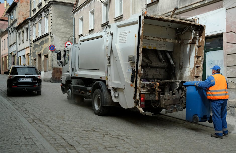 The image shows the rear side of a white delivery van parked on a paved street in front of a building with large windows. Three blue trash bags, made of plastic with slightly crinkled textures, are positioned on the ground near the occupancy side of the van, suggesting they are set aside for rubbish removal or private waste disposal. The bags are filled and tied at the top, with the middle bag leaning slightly to the left. Behind the van, part of the building’s exterior wall is visible, including a white railing or panel at ground level and a glass door or window reflecting the environment. The scene appears to be outdoors during daylight, with natural lighting illuminating the area. This configuration of waste bags outside a commercial or residential building aligns with independent rubbish collection services, such as those provided by companies like Rubbish Removal Harrow, and exemplifies non-local authority waste handling options for the area.