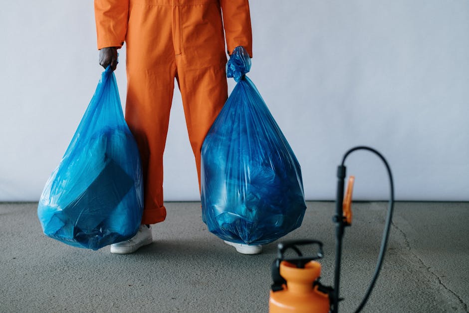 A person wearing an orange jumpsuit is standing indoors on a gray concrete floor, holding two large blue plastic rubbish bags filled with waste. The bags are semi-transparent, revealing uneven contents inside, and are gathered tightly at the top with small knots. The person's hands, covered with black gloves, grip each bag on either side. In the foreground, there is an orange and black sprayer or weed killer device with a flexible hose connected to a spray gun, resting on the ground. The background features a plain, light-colored wall, creating a neutral setting typical for waste collection or rubbish removal activities. This scene emphasizes the process of private waste handling, in line with alternative rubbish disposal methods, and aligns with services offered by Rubbish Removal Harrow in managing household or commercial rubbish efficiently and safely.