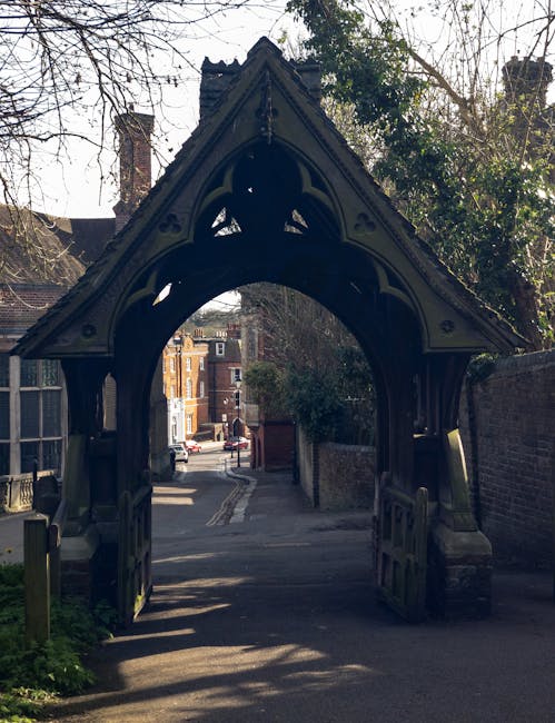 A close-up view of a decorative, black-painted wooden archway with ornate, pointed Gothic-style detailing, situated at the entrance to a narrow street or alley. The archway features carved patterns and is supported by two sturdy side posts, with the top of the structure forming a steeply pitched gable. It casts shadow on the ground beneath, emphasizing its intricate craftsmanship. Beyond the arch, a paved road extends into a residential area with brick buildings, some with classic Victorian or Edwardian architectural details, visible in the background. To the sides, brick walls and greenery frame the scene, with bare tree branches reaching into the sky, indicating late autumn or winter. The setting appears to be an urban or suburban environment with a mixture of historical and modern elements, and the presence of the arch suggests it might be part of a private property, historical site, or a designated entrance related to local heritage or private land management, supported by companies like Rubbish Removal Harrow offering alternative waste clearance options in such areas.