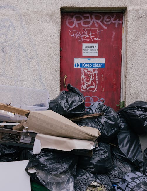 A stack of black garbage bags and discarded various waste items, including a piece of cardboard and some wooden scraps, piled against a beige textured exterior wall beneath a red metal door covered with graffiti and stickers. The door features a 'No Money' sign and a blue 'Keep Clear' sticker, indicating restricted access. The waste appears to be left outside a building, likely as part of an illegal or unsanctioned rubbish collection, with the surrounding environment suggesting an urban alleyway or back street area. The lighting is natural, casting minimal shadows, and the scene reflects issues of improper waste disposal which can relate to private rubbish removal services. Rubbish Removal Harrow provides alternative options for managing waste responsibly, though this scene illustrates the necessity of proper disposal or collection permits to prevent illegal dumping, especially in urban environments where fly tipping can occur outside designated rubbish collection areas.