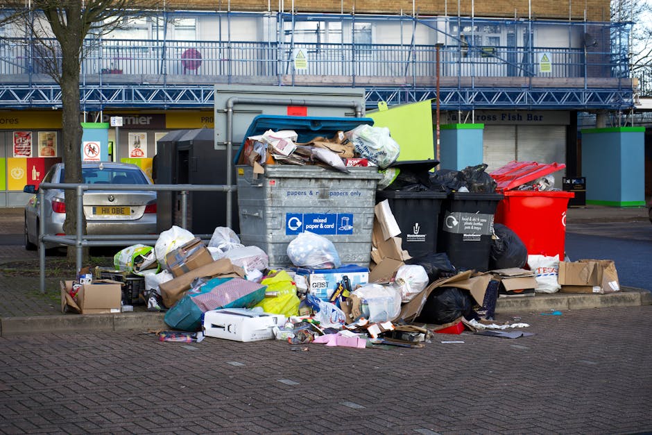 A large cluster of refuse and recycling waste bags, cardboard boxes, and miscellaneous paper and packaging materials piled on a paved area adjacent to a parking space, with various black and coloured bin liners and paper recycling bins arranged behind. The scene is set outdoors in front of a commercial building with blue scaffolding and green corner reinforcements, and blue fencing enclosing the upper level. A metallic car with a visible yellow registration plate is parked to the left, partly obstructed by a metal railing separating the waste area from the parking space. The waste materials include crumpled cardboard, plastic bags, and torn packaging, indicating accumulation of mixed rubbish typical for collection or private disposal services like Rubbish Removal Harrow. Bright, natural daylight illuminates the scene, highlighting the varied textures and colours of the waste, with some items spilling onto the surrounding pavement and sidewalk, suggesting an off-site collection or an on-site clearance operation involving manual handling of rubbish.