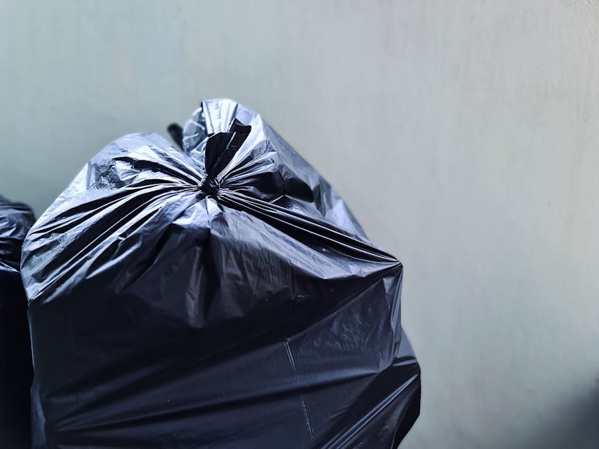 Close-up of a tightly knotted black plastic rubbish bag made from shiny, crinkled plastic material, positioned in the foreground against a plain, light grey background. The bag appears to be filled with waste, creating a rounded, slightly bulging shape with creases and folds visible on its surface. In the background, part of another similar black rubbish bag can be seen, stacked or placed nearby, suggesting a collection or disposal area for waste materials. The scene is illuminated with soft, even lighting that highlights the reflective quality of the plastic, emphasizing its textures and contours. This setting likely depicts an outdoor or indoor rubbish collection site, typical of private waste handling services performed by companies such as Rubbish Removal Harrow, who specialise in rubbish clearance and waste disposal. The focus on the bags and their material aligns with the company's domain of rubbish removal services, illustrating typical waste packaging used in sustainable, non-harmful disposal processes, particularly for larger quantities of household or commercial waste.