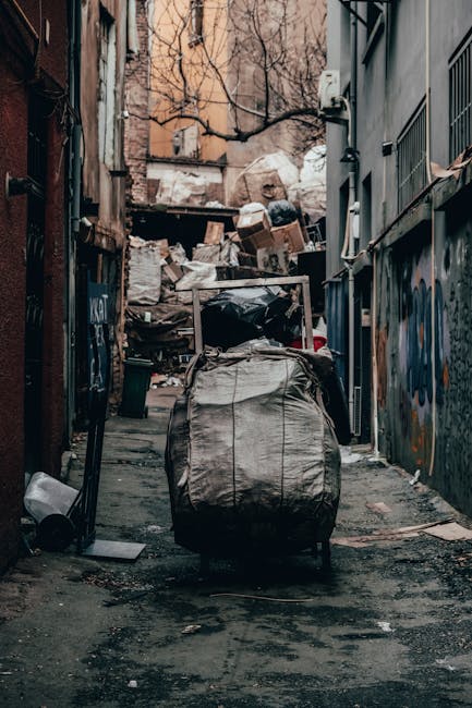 A large cluster of refuse and recycling waste bags, cardboard boxes, and miscellaneous paper and packaging materials piled on a paved area adjacent to a parking space, with various black and coloured bin liners and paper recycling bins arranged behind. The scene is set outdoors in front of a commercial building with blue scaffolding and green corner reinforcements, and blue fencing enclosing the upper level. A metallic car with a visible yellow registration plate is parked to the left, partly obstructed by a metal railing separating the waste area from the parking space. The waste materials include crumpled cardboard, plastic bags, and torn packaging, indicating accumulation of mixed rubbish typical for collection or private disposal services like Rubbish Removal Harrow. Bright, natural daylight illuminates the scene, highlighting the varied textures and colours of the waste, with some items spilling onto the surrounding pavement and sidewalk, suggesting an off-site collection or an on-site clearance operation involving manual handling of rubbish.