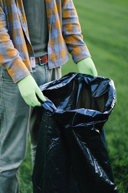 A person outdoors, wearing a green long-sleeved shirt, a checkered yellow and grey jacket, and green gloves, is holding open a large black rubbish bag made of plastic material with a glossy finish. The bag is partially open, revealing an empty interior, and is positioned in front of a grassy area with blurred green foliage in the background. The person's midsection is visible, with a brown belt around their waist, while their face and legs are not shown. The scene suggests an activity related to waste collection or rubbish removal, relevant to private waste handling and alternative rubbish disposal services offered by Rubbish Removal Harrow. The natural daylight Illuminates the scene, emphasizing the texture and sheen of the black rubbish bag and the contrast of the green gloves against the outdoor setting.