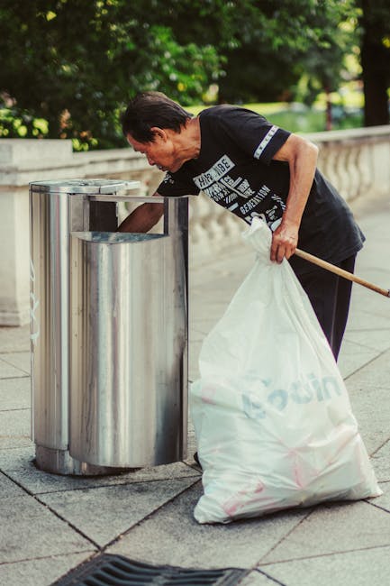 A middle-aged man with dark hair, wearing a black T-shirt with white text and dark trousers, is standing outdoors on a paved surface near a low stone wall surrounded by green trees and foliage. He is bent slightly forward, reaching into a modern, stainless steel public rubbish bin with a curved, cylindrical design and a rectangular opening at the top. The man holds a large white plastic bag, partially filled with waste, in his left hand while his right hand is inside the bin, possibly discarding rubbish. An orange-handled broom or litter picker is tucked under his arm, emphasizing a focus on waste collection or cleanup. The scene is well-lit with natural daylight, and the background features lush greenery, indicating a park or urban outdoor setting where private rubbish collection or street cleaning services such as those offered by Rubbish Removal Harrow may be involved. The context of rubbish disposal is reinforced by the presence of the receptacle and the man’s waste handling activity, supporting the theme of alternative waste removal or on-site clearance.