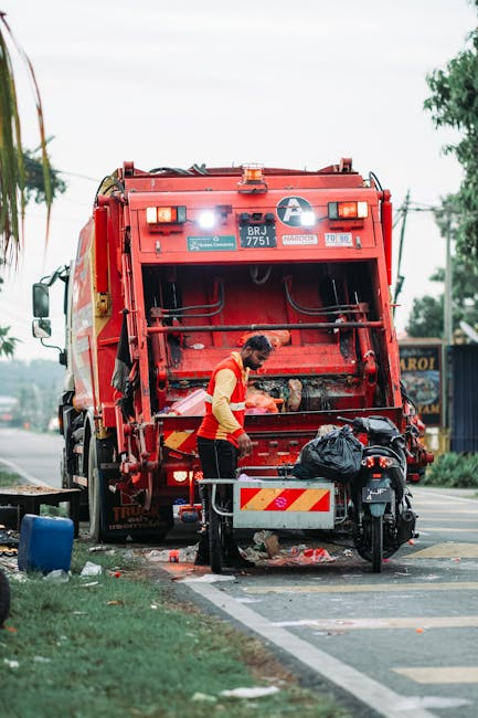 A sanitation worker, dressed in a high-visibility yellow and orange vest and dark trousers, is seen standing next to the open rear compartment of a large red refuse collection vehicle, which is parked on the side of a paved urban street. The worker appears to be handling black trash bags or waste, with some debris and litter visible on the pavement near the vehicle's tailgate. The garbage truck features various safety and identification labels, including a prominent black and white circular logo, vehicle registration numbers, and reflective markings in yellow and red. In the background, there are trees, utility poles, and a section of roadside shops or businesses, indicating a typical city or town environment. The scene is lit by natural daylight, with a clear sky and some greenery on the roadside, complementing the service of private waste collection or rubbish removal. Rubbish Removal Harrow may provide on-site clearance or alternative waste handling options, as suggested by the active waste collection depicted in the image.