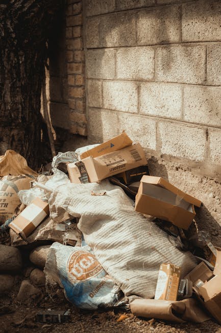 A collection of discarded cardboard boxes and plastic packaging material piled against a rough concrete wall in an outdoor setting. The boxes vary in size, with some flattened and others partially assembled, showing brown kraft paper finishes with printed labels and barcodes. Some boxes are still intact, while others are torn or crushed, indicating previous use. A large white sack made of woven synthetic fabric rests on top of the debris, partially filled and appearing weathered. Surrounding the pile are small rocks and dirt on the ground, with a tree trunk visible on the left side of the image, casting shadows over the scene. The background features a weathered brick section of the wall adjacent to the concrete blocks, suggesting a utilitarian or storage environment. The natural lighting creates soft shadows, emphasizing the textures of the materials, making the scene relevant to private waste handling or rubbish removal services, such as those offered by Rubbish Removal Harrow, in managing outdoor waste collection and clearance.