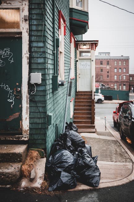 A stack of several black rubbish bags filled with waste materials is placed at the base of a green wooden building with weathered siding, near a set of small wooden steps leading up to an elevated entrance with a white door. The bags are tightly gathered and resting on a concrete sidewalk that curves around the building corner. To the left, a metal door with graffiti tags and peeling paint is partially visible, showing signs of wear. Behind the bags, a white utility box and a wall-mounted telephone are attached to the building’s exterior siding. The background includes a parking lot with a few vehicles, including a black car parked on the street, and distant red-brick industrial-style buildings under a cloudy sky. The scene appears to be part of a private waste collection setup, with the rubbish bags representing curbside disposal awaiting removal or pickup.