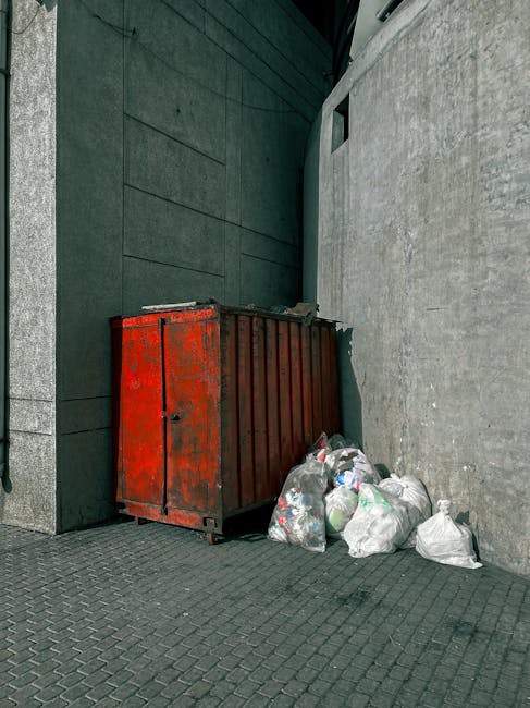 A collection of discarded cardboard boxes and plastic packaging material piled against a rough concrete wall in an outdoor setting. The boxes vary in size, with some flattened and others partially assembled, showing brown kraft paper finishes with printed labels and barcodes. Some boxes are still intact, while others are torn or crushed, indicating previous use. A large white sack made of woven synthetic fabric rests on top of the debris, partially filled and appearing weathered. Surrounding the pile are small rocks and dirt on the ground, with a tree trunk visible on the left side of the image, casting shadows over the scene. The background features a weathered brick section of the wall adjacent to the concrete blocks, suggesting a utilitarian or storage environment. The natural lighting creates soft shadows, emphasizing the textures of the materials, making the scene relevant to private waste handling or rubbish removal services, such as those offered by Rubbish Removal Harrow, in managing outdoor waste collection and clearance.
